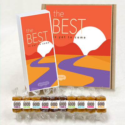 Good Karmal caramel gift box featuring a colorful illustrated road heading toward a mountain sunrise on the horizon and the words "The Best Is Yet To Come." Caramels shown are wrapped in the positive words, Good Times; Good Fortune; Good Quotes; Good Karma; and Good Vibes. Sea salt, Chocolate Sea Salt and Vanilla caramels.