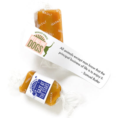 Two caramels are shown: one sea salt caramel with a wrapper featuring an illustration of a dog butt and the words, "Pet All The Dogs, Reduce Stress" and a quote by Samuel Butler that says, "All animals except man know that the principal business of life is to enjoy it." The second caramel is vanilla flavor with a wrapper that says "We are not alone -- Gaze at the Stars"