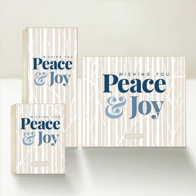 Three holiday-themed Good Karmal caramel gift boxes wishing peace and joy on a winter backdrop of aspens.