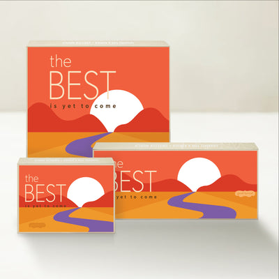 Three holiday-themed Good Karmal caramel gift boxes that say "the best is yet to come" in a hopeful sunny landscape inspired by the road ahead.