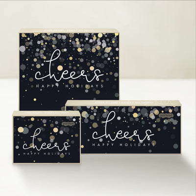 Three holiday-themed Good Karmal caramel gift boxes wishing cheers and happy holidays on a festive background of champagne-like bubbles and confetti.