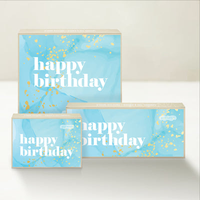 Three Good Karmal caramel gift boxes wishing happy birthday on a blue background with festive confetti-like decorations.