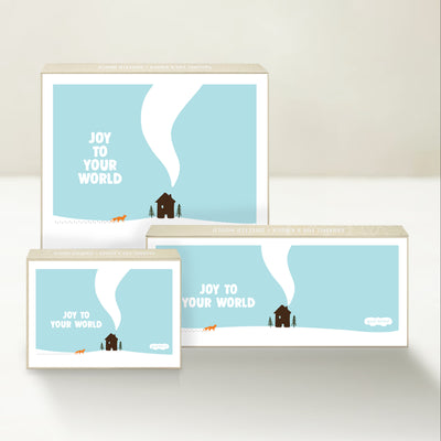 Three Good Karmal caramel gift boxes featuring a whimsical illustration of a fox and a winter house with chimney smoke and "Joy to Your World." 