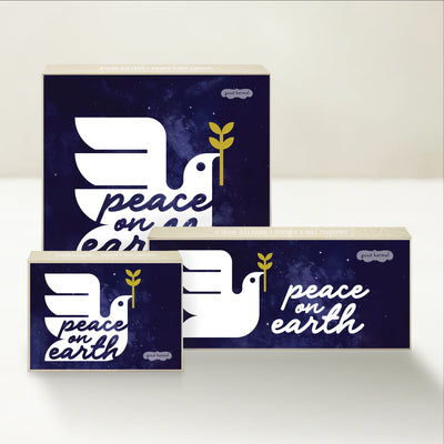 Three holiday-themed Good Karmal caramel gift boxes with an illustration of a dove on a starry backdrop and the words "peace on earth."