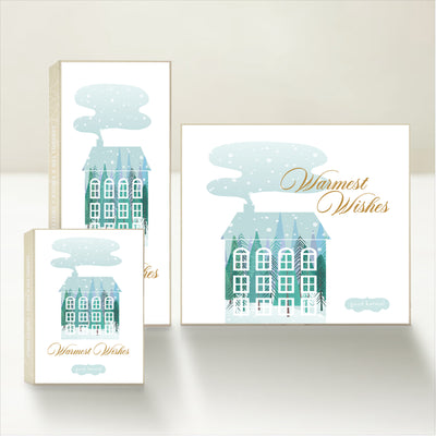 Three holiday-themed Good Karmal caramel gift boxes featuring a whimsical illustration of a winter house with snow and chimney smoke and "Warmest Wishes."