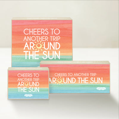 Three Good Karmal caramel gift boxes that say "Cheers to another trip around the sun" on a sunrise-inspired pastel background.